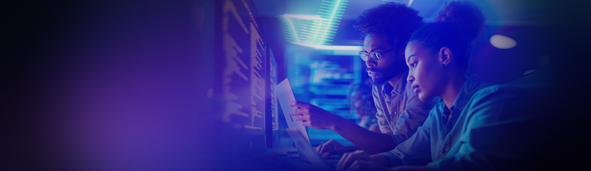 Two professionals analysing data on computer screens in modern office with blue lighting