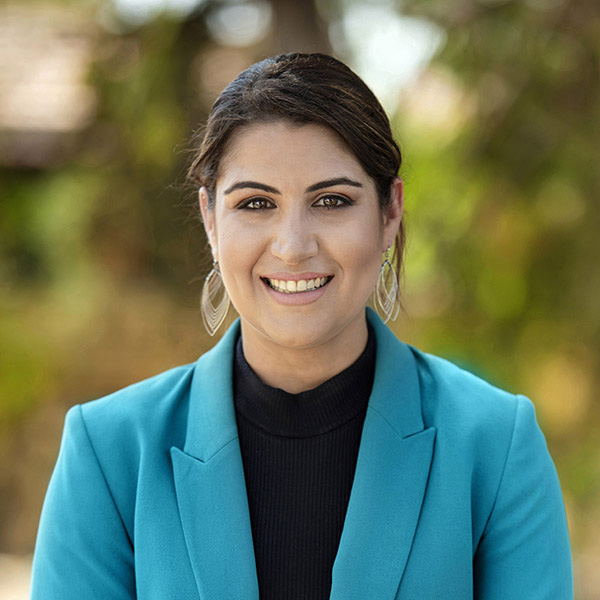 Professional headshot of person smiling in teal blazer outdoors with blurred natural background