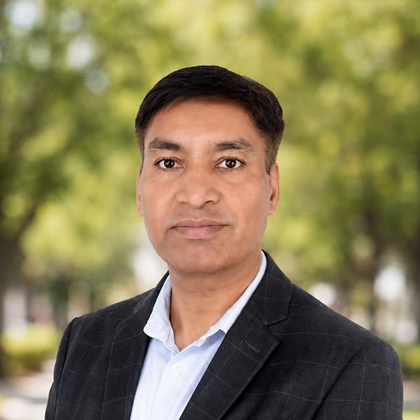 A close-up professional headshot of a person wearing a dark blazer over a light collared shirt, facing the camera with a neutral expression. The background is softly blurred, showing green foliage and an outdoor setting with natural light.