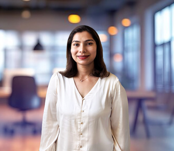 friendly Indian lady wearing light coloured blouse, wearing red lipstick and smiling. The background is an office boardroom, the setting is blurred